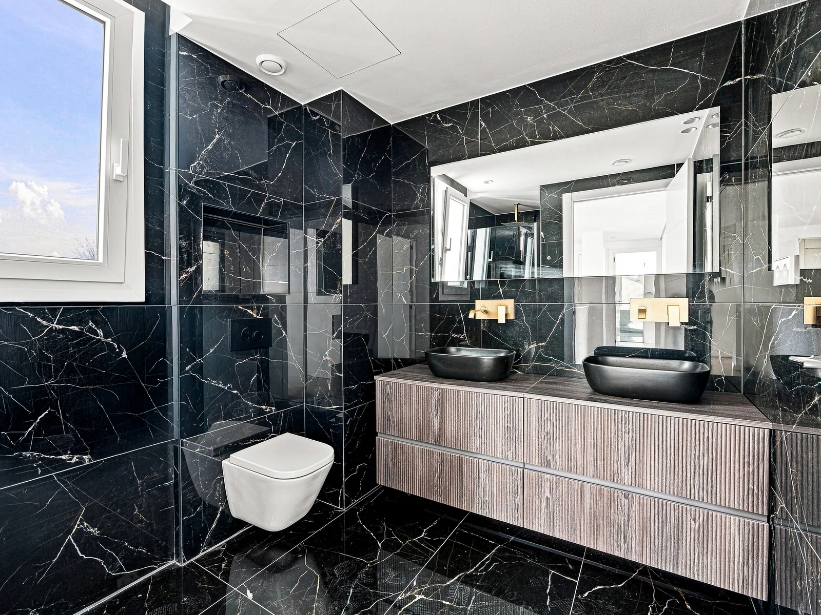 Baño de mármol negro con doble lavabo y grifería dorada, fotografía inmobiliaria de Visoria