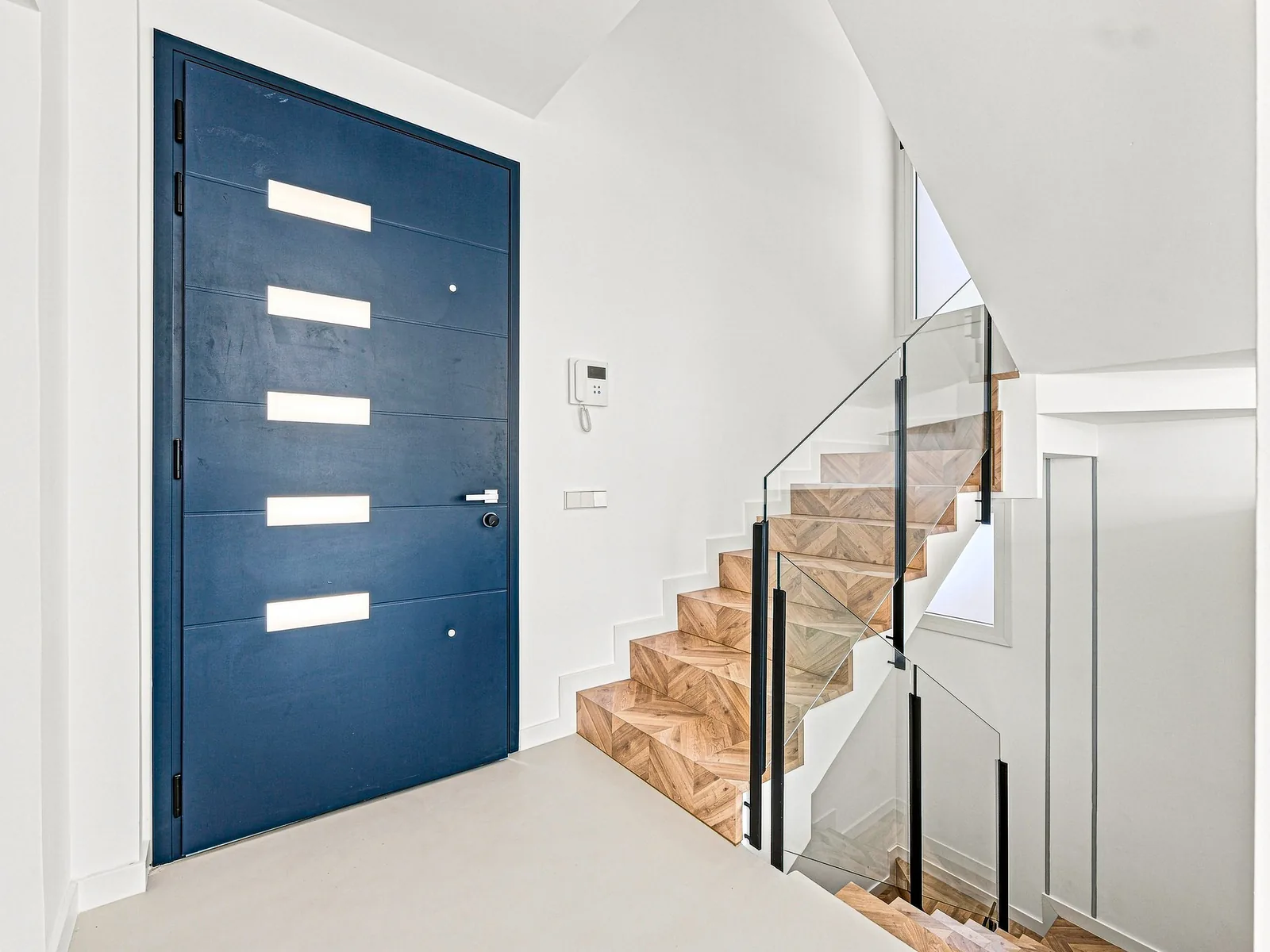 Recibidor con puerta azul y escalera de espiga con barandilla de vidrio, villa en Castelldefels